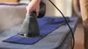 A hand holding a black garment steamer over a blue piece of fabric on an ironing board. Náhľad obrázku produktu