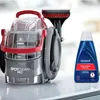 A spot cleaner and a bottle of cleaning solution are displayed on a white surface. Voir les images du produit