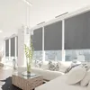 A modern living room with large windows covered by gray roller blinds, furnished with a white sectional sofa and a wooden coffee table. Anteprima dell’immagine prodotto