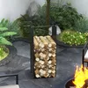 A black metal firewood rack filled with logs is placed in an outdoor garden setting next to a fire pit with flames. Produktbild-Vorschau