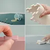 A collage of four images showcasing a baby puzzle mat. The top left shows a teal mat with a spilled cup. The top right shows a hand holding a cream-colored puzzle mat piece. The bottom left shows a hand connecting two pink puzzle mat pieces. The bottom rig Produktbild-Vorschau