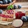 A jar of plum jam with two slices of bread spread with jam, and plums in the background. Produktbild-Vorschau