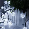 A string of solar-powered LED lights with clear, spherical bulbs is draped over a white picket fence and a potted plant. Produktbild-Vorschau
