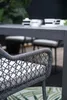 A close-up of a modern outdoor dining chair with a grey rope weave and a light-colored cushion, positioned in front of a dark grey outdoor table and chairs. A tropical-themed rug is visible on the floor. Produktbild-Vorschau