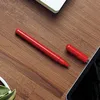 A red pen and its cap are placed on a wooden desk next to a tablet and a notebook. Produktbild-Vorschau