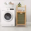 A laundry room scene with a washing machine, a bamboo shelving unit with towels and bottles, and a basket of folded towels on top of the washing machine. Produktbild-Vorschau
