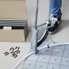 A person is using a tool to attach a white pipe to a grid on the floor, with a box of fasteners in the background. Náhľad obrázku produktu