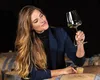 A woman is holding a wine glass and looking at it, with wine barrels in the background. Produktbild-Vorschau