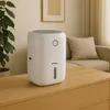 A white dehumidifier is placed on a wooden table in a living room setting, with a plant and a sofa in the background. Podgląd zdjęcia produktu