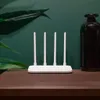 A white router with four antennas is placed on a brown surface in front of a green wall. A dark bottle and green leaves are also visible in the background. Podgląd zdjęcia produktu
