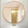 A round mirror with a gold frame is mounted on a marble wall above a sink with gold faucets. The mirror reflects a bathroom interior with beige tiles, a shower head, and a towel rack. Náhľad obrázku produktu