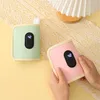 Two portable bottle warmers, one green and one pink, are shown on a yellow and white lace-covered surface. A person's hands are holding the pink bottle warmer. Produktbild-Vorschau