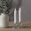 Two LED candles in glass holders on a wooden table, with a vase and greenery in the background. Produktbild-Vorschau