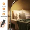 A floor lamp is shown next to a bed with pillows and a blanket. A remote control is held in a hand in the foreground. Produktbild-Vorschau