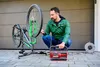A man is using a portable air compressor to inflate a bicycle tire. The bicycle is mounted on a stand. A coiled extension cord is visible in the background. Voir les images du produit