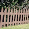 A brown picket fence in front of green bushes and grass. Náhľad obrázku produktu