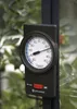A thermometer with a black casing and a white dial is mounted on a dark pole, with a blurred green background of plants. Produktbild-Vorschau