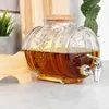 A glass beverage dispenser with a spigot and cork lid, filled with amber liquid, sits on a countertop. A wooden stand is to the left, and plants are in the background. Produktbild-Vorschau