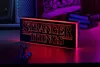 A Stranger Things logo light is displayed on a desk with a small figurine in front of it, illuminated by red light. Produktbild-Vorschau