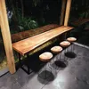 A long wooden bar table with four stools in an outdoor setting with lush greenery visible through a large window. Produktbild-Vorschau