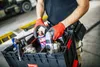 A person wearing red gloves is holding a black toolbox filled with various tools and spray cans. Náhled obrázku produktu