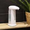 A white automatic soap dispenser sits on a wooden shelf in front of a black tiled wall, with a green plant in a woven basket to the right. Produktbild-Vorschau