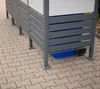 A close-up view of a gray metal trash can enclosure with horizontal slats, showing a blue bin and a wheel underneath. Náhled obrázku produktu