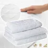 A hand is shown touching a folded white mattress protector, with a close-up inset of its quilted texture. Three folded mattress protectors are stacked in the foreground. Produktbild-Vorschau