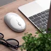 A silver Satechi M1 Bluetooth wireless mouse is placed on a wooden desk next to a laptop and a small green plant. A pair of glasses is also visible in the foreground. Anteprima dell’immagine prodotto