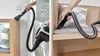 A person is using a vacuum cleaner attachment to clean a wooden surface and a sofa cushion. Voir les images du produit