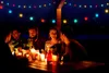 A group of friends are enjoying a festive outdoor dinner party at night, illuminated by colorful string lights and candles. Produktbild-Vorschau
