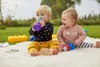 Two babies are sitting on a white blanket outdoors, with toys and bottles around them. One baby is drinking from a purple sippy cup, and the other is looking to the side. Voir les images du produit