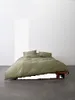 A bed with green bedding is shown in a room with a white wall and gray floor. Produktbild-Vorschau