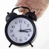 A hand is holding a black retro analog alarm clock. Voir les images du produit