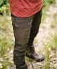 A person wearing outdoor clothing, including pants and boots, standing in a natural, wooded environment. Produktbild-Vorschau