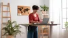 A woman is working on a laptop at a standing desk in a bright, modern office space. The room is decorated with plants and a world map on the wall. Produktbild-Vorschau