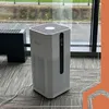 An air purifier is placed on a carpeted floor next to a window, with a black object to its left. Náhled obrázku produktu
