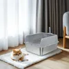 A cat is lying on a fluffy white mat next to a stainless steel litter box with a scoop. Produktbild-Vorschau