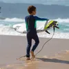 A young boy in a wetsuit walks on a beach carrying a surfboard. Produktbild-Vorschau