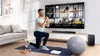A man is doing a lunge exercise in front of a large TV screen displaying a fitness video. Náhled obrázku produktu