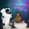 A child is sitting on a bed, looking at a projector that is casting a starry sky onto the ceiling. A remote control is placed on the bed. Produktbild-Vorschau
