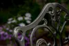 Close-up of a decorative metal chair back with blurred flowers in the background. Produktbild-Vorschau