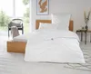 A bedroom scene with a bed made up with white bedding, a chair with a blanket, and a small side table. Produktbild-Vorschau