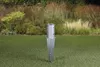A metal ground post anchor is shown in a grassy yard with a blurred background of plants and grasses. Produktbild-Vorschau