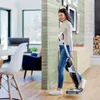 A woman is vacuuming a wooden floor with a BISSELL Crosswave vacuum cleaner in a modern living room. Voir les images du produit