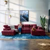 A woman sits on a large U-shaped sofa, using a laptop. A dog lies on the sofa next to her. The room is decorated with a large piece of art and a tall plant. Produktbild-Vorschau