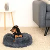 A small dog is resting in a plush donut bed on a carpeted floor next to a sofa and a side table. Anteprima dell’immagine prodotto
