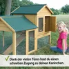 A young girl is sitting on the grass next to a wooden rabbit hutch with an open door. Produktbild-Vorschau