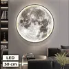 A large, illuminated moon-shaped wall lamp is mounted on a gray wall above a black leather sofa and a modern side table. Books are stacked on the side table, and a vase with dried flowers is placed on the sofa. A bookshelf with decorative items is visible  Náhled obrázku produktu
