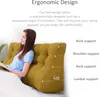 A woman is sitting up in bed, leaning against a large wedge pillow, reading a book. The pillow is yellow and has multiple support areas labeled. Produktbild-Vorschau
